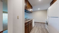 This single-floor unit offers a practical and functional layout with distinct living spaces. The kitchen is designed in a galley style, featuring dark wood cabinetry with gold hardware extending both above and below the countertops, laminate counters, a double-basin sink, a white electric range with overhead ventilation, a refrigerator, and a dishwasher. Light wood-look vinyl flooring runs throughout the kitchen, which also includes a closet for additional storage. A peninsula with a built-in sink and matching countertop separates the kitchen from the adjacent living area, adding a degree of openness to the layout.

The living area is generously sized with neutral carpet and is anchored by a large sliding glass door fitted with vertical blinds, allowing natural light to filter in and offering views of surrounding trees. A wall-mounted air conditioning unit and baseboard heating provide year-round climate control. The bedroom is carpeted and features a window with vertical blinds and a baseboard heater beneath it, providing a quiet, functional space. The bathroom includes a tiled floor in a classic mosaic pattern, a full tub and shower combination with a curved curtain rod, a toilet, and dark wood vanity cabinetry with a light countertop.