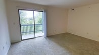 This single-floor space features a clean, open living area finished with wall-to-wall neutral carpeting that adds a sense of warmth and comfort underfoot. The room is kept bright by a large sliding glass door fitted with vertical blinds, which serves as both a generous source of natural light and a direct access point to a private balcony. The balcony, enclosed by a classic railing, overlooks a lush, green landscape with mature trees visible in the distance, providing a peaceful outdoor retreat. The neutral-toned walls complement the light carpeting, creating a cohesive and understated aesthetic throughout the space. A ceiling vent indicates a central HVAC system for year-round climate control. The layout feels open and versatile, offering ample room for a variety of furniture arrangements to suit different lifestyle needs. Overall, the space presents a functional, well-lit environment with a comfortable connection to the outdoors through its balcony access and expansive glazing.