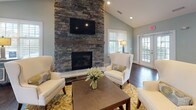 Located at 1100 Deer Hunter Dr in Garner, NC, this single-story community clubhouse offers an open, well-appointed gathering space designed for comfort and functionality. The main lounge area serves as the focal point of the space, anchored by a floor-to-ceiling stacked stone fireplace with a wood mantel and wall-mounted television. Vaulted ceilings enhance the sense of openness, while dark hardwood floors run throughout, providing a warm contrast to the soft sage-green walls. Multiple sets of French doors allow generous natural light to flow in and offer views of the surrounding community.

The kitchen area is equipped with stainless steel appliances, including a full-size refrigerator, dishwasher, and microwave. White upper cabinetry pairs with granite countertops along the perimeter, while a curved granite-topped bar with woven barstool seating serves as a natural divider between the kitchen and lounge. A casual high-top dining area near the French doors provides an additional seating option. Recessed lighting and vaulted ceilings carry throughout the space, maintaining a consistent, airy ambiance. The overall design balances practical amenities with an inviting, residential aesthetic suited for community gatherings.