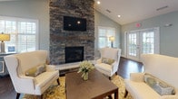 Located at 1100 Deer Hunter Dr in Garner, NC, this single-story community clubhouse offers an open, well-appointed gathering space designed for comfort and functionality. The main lounge area serves as the focal point of the space, anchored by a floor-to-ceiling stacked stone fireplace with a wood mantel and wall-mounted television. Vaulted ceilings enhance the sense of openness, while dark hardwood floors run throughout, providing a warm contrast to the soft sage-green walls. Multiple sets of French doors allow generous natural light to flow in and offer views of the surrounding community.

The kitchen area is equipped with stainless steel appliances, including a full-size refrigerator, dishwasher, and microwave. White upper cabinetry pairs with granite countertops along the perimeter, while a curved granite-topped bar with woven barstool seating serves as a natural divider between the kitchen and lounge. A casual high-top dining area near the French doors provides an additional seating option. Recessed lighting and vaulted ceilings carry throughout the space, maintaining a consistent, airy ambiance. The overall design balances practical amenities with an inviting, residential aesthetic suited for community gatherings.