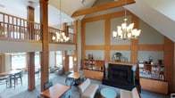 This amenity space is designed to offer residents a refined and functional environment across two distinct areas. The grand lounge immediately commands attention with its soaring double-height ceilings, exposed wood beam accents, and warm-toned wood paneling that frame the room with architectural elegance. A striking floor-to-ceiling fireplace with a dark stone surround serves as the focal point, flanked by built-in bookshelves and cabinetry. Stylish multi-light chandeliers illuminate the space, which is furnished with comfortable seating arrangements and dining tables ideal for socializing or quiet relaxation. A mezzanine-level overlook with wooden railings adds to the vertical drama of the room, while large windows invite natural light and views of the surrounding greenery.

The fitness center is a comprehensive, well-lit facility outfitted with a full suite of Life Fitness strength training machines and a dedicated cardio zone featuring rows of treadmills and elliptical trainers equipped with individual screens. Light wood-look flooring, mirrored walls, and a vibrant teal accent wall create a bright and motivating atmosphere. A water station and dumbbell rack are conveniently accessible, and additional rooms off the main floor provide private changing or storage space. The facility is thoughtfully designed to support a complete workout experience.