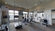 This fitness center in Dallas, Texas, offers a well-appointed space designed to support a range of cardio-focused workouts. The room is finished with neutral-toned walls accented by a distinctive navy blue horizontal stripe, giving the space a clean, modern aesthetic. Durable patterned carpet flooring covers the workout area, providing both comfort and traction underfoot.

Large multi-pane windows line the walls, flooding the interior with abundant natural light while offering views of the surrounding outdoor environment. Ceiling fans are mounted throughout, ensuring consistent air circulation during workouts. Recessed lighting supplements the natural light, keeping the space well-illuminated at all times.

The fitness center is equipped with a variety of cardio machines, including treadmills, elliptical trainers, a recumbent bike, and stationary cycles. A wall-mounted flat-screen television provides entertainment during exercise sessions. A large framed mirror along one wall visually expands the space and allows users to monitor their form. The overall ambiance is bright, functional, and inviting, making it a practical and comfortable environment for daily fitness routines.
