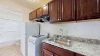 This single-floor unit offers a practical and well-organized layout across several distinct living areas. The kitchen is outfitted with rich, dark-stained wood cabinetry featuring raised-panel doors and brushed nickel hardware, complemented by granite-look countertops and a stainless steel sink. White appliances — including a coil-burner range, a range hood, and a full-size refrigerator — complete the functional cooking space. The kitchen opens directly to a carpeted living area, creating a connected and efficient flow. Neutral white walls and ceiling-mounted lighting keep the shared space feeling bright and clean. A carpeted bedroom offers a quiet retreat, featuring a window that admits natural light and a door providing convenient exterior access. Sliding glass doors in one of the rooms open to a view of lush green outdoor surroundings, allowing generous natural light to fill the space. Baseboard heating is present throughout the carpeted areas, ensuring year-round comfort. The bathroom is finished with tile flooring in a warm beige tone and includes a full tub-shower surround with built-in shelving, a standard toilet, and a wood-toned vanity with a white countertop and mirror cabinet above. Overall, the unit presents a clean, move-in-ready environment with a straightforward, functional aesthetic.