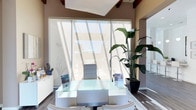 This contemporary single-floor professional office space makes a strong first impression with its striking architectural design and bright, welcoming atmosphere. The reception area serves as the focal point of the space, anchored by a sleek white glass-topped desk that reflects the clean, modern aesthetic carried throughout. Exposed wooden ceiling beams add a warm, organic contrast to the otherwise crisp interior, creating a balanced blend of warmth and sophistication.

Floor-to-ceiling windows dominate one wall, flooding the space with abundant natural light and casting dramatic diagonal shadows across the hardwood flooring — a dynamic visual feature that shifts throughout the day. Sheer window treatments allow light to diffuse softly while maintaining a sense of privacy. The tall ceilings enhance the open, airy feel of the space, giving the interior a sense of volume and grandeur.

Tasteful touches of greenery, including a large tropical Bird of Paradise plant, bring life and color to the neutral, earth-toned palette of warm beige and taupe walls. The overall ambiance is professional yet inviting, thoughtfully designed to accommodate both staff and visiting clients. Informational displays and literature holders near the reception desk further reinforce the space's functional, client-forward purpose.