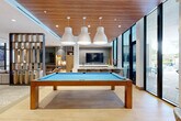 Located at 776 Riverside Dr in Coral Springs, Florida, this Club Room is a thoughtfully designed single-floor amenity space offering a generous and versatile layout. The interior showcases a contemporary aesthetic with a warm wood-paneled ceiling in the central entertainment zone, complemented by light hardwood flooring that flows throughout. A full-size pool table anchors the main area beneath a trio of oversized white pendant lights, creating a striking focal point. A built-in media wall with a large flat-screen TV and wet bar serves as an entertainment hub, ideal for gatherings. Floor-to-ceiling glass walls and doors flood the space with abundant natural light and provide views of the outdoor surroundings, seamlessly connecting interior and exterior. A wall of open wood shelving adds architectural interest and display functionality, while a decorative lattice-style partition elegantly defines distinct zones within the open plan. Multiple seating areas are distributed throughout, including a lounge with modish swivel chairs, a cozy sofa nook with built-in lit shelving, and an entry foyer featuring a tufted bench and vibrant large-scale floral artwork. Patterned area rugs define each zone, adding texture and visual warmth. The overall ambiance is modern, bright, and inviting — purpose-built for social interaction and relaxation.
