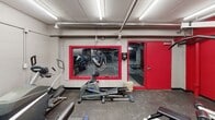 Located at 1015 SE Eighth St in Minneapolis, MN, this fitness center is a well-equipped, single-floor exercise facility designed to support a well-rounded workout routine. The space features a striking interior defined by bold red accent walls set against light gray painted concrete block, giving the room a energetic and motivating atmosphere. Overhead fluorescent strip lighting provides consistent, bright illumination throughout the space, while exposed ductwork and piping along the ceiling contribute to the facility's utilitarian, industrial character. Large wall-mounted mirrors line portions of the room, amplifying the sense of space and allowing users to monitor their form during exercise. The fitness center is outfitted with a variety of cardio equipment, including treadmills, elliptical trainers, and a stationary bike. A dedicated strength training area features a multi-station cable and weight machine, as well as a flat bench for free weight exercises. A full rack of dumbbells in graduated sizes lines one wall, catering to a range of fitness levels. A wall-mounted flat-screen television is positioned for easy viewing from the cardio area. A large window with a bold red frame overlooks an adjacent parking structure, and a red steel door provides direct access to the building. Rubber flooring throughout the space offers durability and safety underfoot.
