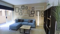 This single-floor space centers around a well-appointed living area designed for comfort and visual interest. The room features a spacious blue-gray sectional sofa with a chaise, accented by striped and solid throw pillows in brown and tan tones. A light-toned rectangular coffee table sits in front of the sofa, providing a functional surface. A standout design feature is the gallery wall, which displays an eclectic arrangement of empty picture frames in varying sizes, shapes, and finishes — including black, white, natural wood, and ornate styles — creating a striking focal point against the warm beige walls. A tall, cylindrical modern floor lamp in the corner emits a soft, ambient glow, contributing to the room's relaxed atmosphere. A dark bookshelf along the right wall adds storage and depth to the space. The flooring is carpeted in a neutral tone, complementing the overall warm and cohesive color palette. The ceiling is smooth and white, helping to brighten the interior. The space conveys a creative, casual ambiance with a personalized touch through its distinctive wall decor.