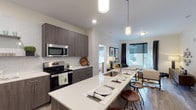 The Edge on 4th presents a thoughtfully designed single-floor residence that combines contemporary style with functional living. The open-concept kitchen is a standout feature, showcasing dark wood-grain cabinetry, quartz countertops, a white subway tile backsplash, and a full suite of stainless steel appliances including a French door refrigerator and electric range. A generously sized island with bar seating and pendant lighting serves as the centerpiece, flowing seamlessly into the living area, which is brightened by large windows with urban views. The living room offers a comfortable, well-lit retreat with hardwood-style flooring throughout the common areas. The residence includes multiple bedrooms, each carpeted and equipped with ceiling fans and windows with blackout curtains. The primary bedroom connects to an en-suite bathroom featuring a glass-enclosed shower, wood-tone vanity with quartz countertop, and direct access to a walk-in closet. A second full bathroom includes a shower-tub combo and a matching vanity, with a stacked washer and dryer conveniently located just steps away. The neutral color palette and modern finishes throughout create a cohesive and contemporary ambiance.