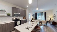 The Edge on 4th presents a thoughtfully designed single-floor residence that combines contemporary style with functional living. The open-concept kitchen is a standout feature, showcasing dark wood-grain cabinetry, quartz countertops, a white subway tile backsplash, and a full suite of stainless steel appliances including a French door refrigerator and electric range. A generously sized island with bar seating and pendant lighting serves as the centerpiece, flowing seamlessly into the living area, which is brightened by large windows with urban views. The living room offers a comfortable, well-lit retreat with hardwood-style flooring throughout the common areas. The residence includes multiple bedrooms, each carpeted and equipped with ceiling fans and windows with blackout curtains. The primary bedroom connects to an en-suite bathroom featuring a glass-enclosed shower, wood-tone vanity with quartz countertop, and direct access to a walk-in closet. A second full bathroom includes a shower-tub combo and a matching vanity, with a stacked washer and dryer conveniently located just steps away. The neutral color palette and modern finishes throughout create a cohesive and contemporary ambiance.