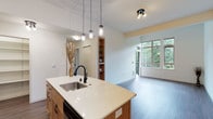 This well-appointed single-floor residence offers a seamless open-concept layout that connects the kitchen and living area in a clean, modern aesthetic. The kitchen is anchored by a central island with a crisp white quartz countertop, an undermount stainless steel sink, and a matte black faucet that adds a contemporary contrast. Warm wood cabinetry brings a natural warmth to the space, while three elegant pendant lights suspended above the island provide focused task lighting and a refined visual accent. The kitchen flows effortlessly into the living area, which is bathed in natural light from large floor-to-ceiling windows that frame views of surrounding mature greenery, creating a serene, nature-connected atmosphere. A sliding glass door leads to an outdoor balcony, further extending the sense of openness. The living area features smooth hardwood-style flooring that runs throughout, contributing to a cohesive and polished look. A built-in wooden shelving unit adds both functional storage and a warm design detail between rooms. The high, smooth ceilings enhance the feeling of spaciousness throughout. Overall, the space strikes a balance between modern sophistication and comfortable, livable design.