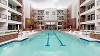 This outdoor amenity space is centered around a generously sized lap pool with clearly marked swim lanes, finished in crisp aqua-blue tile and surrounded by a wide, light-toned concrete pool deck. White modern sun loungers are arranged along both sides of the pool, offering ample space for relaxation. The pool is framed on multiple sides by a contemporary multi-story building featuring a striking combination of red brick and light stucco facades, large energy-efficient windows, and private balconies on upper floors. Floor-to-ceiling glazed panels at the ground level provide visual connectivity between interior common areas and the outdoor deck. A shaded seating area near the building entrance features round tables with vibrant blue patio umbrellas and green sling chairs, ideal for outdoor dining or socializing. On one side of the courtyard, a dedicated outdoor grill station is built into a brick surround beneath a pergola-style overhead structure, complete with a full-size stainless steel gas grill and utility sink. Planters and seasonal trees add organic warmth to the hardscaped environment, while the enclosed courtyard design fosters a sense of privacy and community throughout this well-appointed outdoor retreat.