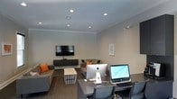 This single-floor space serves a dual purpose, seamlessly integrating a comfortable lounge area with a functional workstation zone. The room features neutral gray walls complemented by crisp white crown molding, lending a clean and polished aesthetic throughout. Recessed lighting is evenly distributed across the ceiling, ensuring consistent illumination across both zones.

The lounge area is furnished with contemporary gray sofas accented by vibrant orange throw pillows, adding a pop of color to the otherwise neutral palette. A wall-mounted flat-screen television sits atop a low-profile media console, making this area well-suited for presentations or casual viewing. A wire-frame coffee table serves as a central accent piece between the seating arrangements.

Transitioning to the workspace side, the area is equipped with multiple desktop computers, including an Apple iMac and an additional all-in-one unit, set upon a dark-toned workstation desk with ergonomic seating. A framed artwork piece on the wall adds a subtle decorative touch. The overall ambiance is professional yet welcoming, reflecting a versatile communal space designed to accommodate both collaboration and productivity.