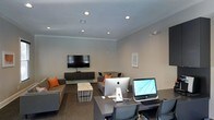 This single-floor space serves a dual purpose, seamlessly integrating a comfortable lounge area with a functional workstation zone. The room features neutral gray walls complemented by crisp white crown molding, lending a clean and polished aesthetic throughout. Recessed lighting is evenly distributed across the ceiling, ensuring consistent illumination across both zones.

The lounge area is furnished with contemporary gray sofas accented by vibrant orange throw pillows, adding a pop of color to the otherwise neutral palette. A wall-mounted flat-screen television sits atop a low-profile media console, making this area well-suited for presentations or casual viewing. A wire-frame coffee table serves as a central accent piece between the seating arrangements.

Transitioning to the workspace side, the area is equipped with multiple desktop computers, including an Apple iMac and an additional all-in-one unit, set upon a dark-toned workstation desk with ergonomic seating. A framed artwork piece on the wall adds a subtle decorative touch. The overall ambiance is professional yet welcoming, reflecting a versatile communal space designed to accommodate both collaboration and productivity.