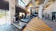 Located at 750 Thornton Way in Alexandria, Virginia, this two-story fitness center is a well-appointed facility designed to support a comprehensive range of workout routines. The space is defined by soaring double-height ceilings, abundant natural light from large windows, and a refined mix of hardwood flooring and patterned carpet that delineates distinct training zones throughout.

The lower level houses an extensive cardio area lined with treadmills, ellipticals, stair climbers, and stationary bikes, all positioned along windowed walls that offer views of the surrounding exterior. Dark brick accent walls and warm wood paneling contrast with crisp white structural columns, lending the space a modern yet inviting atmosphere. A central functional training corridor features a dedicated hardwood sprint lane and a built-in wall-mounted display, ideal for dynamic workouts.

A sleek staircase with glass railings and brushed steel accents connects the lower level to the upper floor, which overlooks the cardio zone below and hosts additional strength training equipment, cable machines, and a free weight area with a full dumbbell rack. Pendant lighting and recessed linear fixtures illuminate both levels, contributing to a clean, contemporary aesthetic. Together, the two floors form a cohesive, fully equipped fitness environment suited to a wide variety of training needs.