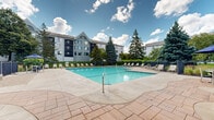 Located at 1180 Cushing Cir in Saint Paul, Minnesota, this expansive outdoor amenity space offers a well-appointed patio and grounds designed for both relaxation and recreation. The centerpiece of the outdoor area is a generously sized swimming pool surrounded by a stamped concrete deck lined with lounge chairs, perfect for enjoying sunny Minnesota days beneath open skies. Adjacent to the pool, a gated entry with manicured evergreen shrubs provides a welcoming transition from the surrounding grounds.

The landscaping throughout is thoughtfully maintained, featuring a mix of mature deciduous trees, blue spruce, and ornamental plantings bordered by natural stone edging. Winding paved pathways meander across lush, rolling lawns, connecting the various recreational areas and building entrances. A dedicated lawn games area includes a cornhole setup and a bocce ball court with brick-paver borders, offering casual outdoor entertainment. A shaded patio with brick pavers, seating, and a large shade tree provides a quiet retreat for gathering.

The multi-building backdrop features a distinctive multi-story architectural design with gabled rooflines and large windows. The overall ambiance is park-like, combining active amenities with serene green spaces that create an inviting and well-rounded outdoor experience.