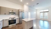Located at 1720 Nick Nuccio Parkway in Tampa's vibrant Ybor City neighborhood, this modern 2-bedroom, 2-bathroom residence offers a thoughtfully designed single-floor layout with contemporary finishes throughout. The open-concept living and kitchen area serves as the heart of the home, featuring soaring ceilings, recessed lighting, and rich wood-look flooring that flows seamlessly throughout the main living spaces. The well-appointed kitchen is equipped with a full suite of stainless steel appliances, including a French door refrigerator, electric range, built-in microwave, and dishwasher. Sleek two-tone cabinetry, a chevron-patterned tile backsplash, white quartz countertops, and a central island with additional prep space complete the culinary space. The living area is bathed in natural light from French doors with transom windows above, opening to an outdoor view. The primary bedroom features soft carpeting, elevated ceilings, a stylish sliding barn door leading to a private en-suite bathroom, and a spacious walk-in closet with wire shelving. The second bedroom also offers plush carpeting and large windows. Both bathrooms are finished with modern vanities featuring wood-toned cabinetry and framed mirrors, with the primary bath showcasing a walk-in glass shower with floor-to-ceiling white tile.