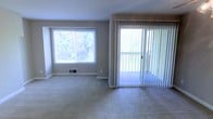 This single-floor space is designed with comfort and natural light in mind. The living area is finished with wall-to-wall carpeting in a neutral tone, creating a warm and inviting atmosphere throughout. A large picture window dominates one wall, flooding the room with abundant natural light while framing picturesque views of the surrounding greenery. Adjacent to the window, a full-length sliding glass door fitted with vertical blinds provides direct access to an outdoor area, further enhancing the connection between the interior and the natural environment outside. The walls are painted in a soft, neutral gray, contributing to a clean and understated aesthetic that complements a variety of furnishing styles. A floor-level air vent is integrated into the baseboard, indicating a central heating and cooling system for year-round climate comfort. The overall layout is open and unobstructed, offering flexible arrangement possibilities. The combination of plush carpeting, ample natural light, and outdoor access makes this space well-suited for relaxed everyday living.