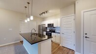 Located at 291 Graceland Blvd in Columbus, Ohio, this single-floor residence offers a well-designed open-concept layout with modern finishes. The kitchen serves as the heart of the home, showcasing a large center island with a dark granite countertop and an integrated sink, complemented by elegant pendant lighting overhead. White shaker-style cabinetry lines the walls, providing ample storage, while a subway tile backsplash and a full suite of stainless steel appliances—including a refrigerator, electric range, and microwave—complete the space. Warm hardwood-style flooring flows throughout the kitchen and living areas, creating a cohesive and inviting atmosphere. The living area transitions seamlessly from the kitchen and features plush carpeting, with sliding glass doors that allow natural light to pour in and provide access to an outdoor view. A secondary bedroom mirrors this comfort with soft carpeting and a bright window overlooking the exterior. The bathroom is appointed with dark granite countertops, white cabinetry, and hardwood-style flooring, and connects directly to a spacious walk-in closet outfitted with built-in shelving and hanging rods—offering practical and generous storage. The overall design balances contemporary aesthetics with functional living.