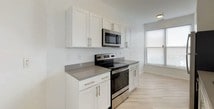 This well-appointed single-floor residence offers a clean, modern aesthetic throughout, characterized by crisp white walls, light-toned luxury vinyl plank flooring in the main living areas, and an abundance of natural light. The open-concept living room is a standout space, featuring a sleek fireplace flanked by large windows and a sliding glass door that leads to a private outdoor balcony, filling the room with natural light and outdoor views. The kitchen is thoughtfully designed with white shaker-style cabinetry, grey countertops, and a full suite of stainless steel appliances including a refrigerator, electric range, and built-in microwave. A peninsula counter separates the kitchen from the living area, creating a practical and social layout. The bedrooms are comfortably finished with plush wall-to-wall carpeting and generous windows that invite natural light. The primary bathroom features a full tub-shower combination with white tile surround, a wide vanity with ample counter space, and a large mirror. A second bathroom offers a single vanity with under-sink cabinetry and tile flooring, maintaining the same clean, neutral palette found throughout the residence. The overall ambiance is bright, fresh, and move-in ready.
