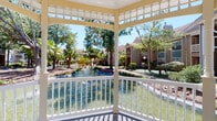 Gazebo 1 is a single-story amenity space combining a charming outdoor gazebo with a fully equipped fitness center. The gazebo features classic white-painted woodwork with decorative lattice detailing and a traditional railing, overlooking a serene pond surrounded by mature tropical trees and manicured landscaping. The tranquil waterfront setting provides a peaceful retreat within the broader community.

The fitness center is bright and modern, featuring neutral gray walls, recessed lighting, and ceiling fans that maintain a comfortable environment throughout. Floor-to-ceiling mirrors line the walls, visually expanding the space and supporting proper form during workouts. Large windows and glass sliding doors flood the interior with natural light while offering views of the outdoor pool area.

The facility is comprehensively outfitted with multiple treadmills, ellipticals, and stationary bikes featuring integrated screens for cardio training. Strength equipment includes multi-function cable machines, adjustable benches, a full rack of dumbbells, and a diverse selection of medicine balls displayed on dedicated racks. Organized open shelving holds rolled towels and accessories, keeping the space tidy and functional. A wall-mounted television adds entertainment during workouts, enhancing the overall fitness experience.