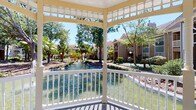 Gazebo 1 is a single-story amenity space combining a charming outdoor gazebo with a fully equipped fitness center. The gazebo features classic white-painted woodwork with decorative lattice detailing and a traditional railing, overlooking a serene pond surrounded by mature tropical trees and manicured landscaping. The tranquil waterfront setting provides a peaceful retreat within the broader community.

The fitness center is bright and modern, featuring neutral gray walls, recessed lighting, and ceiling fans that maintain a comfortable environment throughout. Floor-to-ceiling mirrors line the walls, visually expanding the space and supporting proper form during workouts. Large windows and glass sliding doors flood the interior with natural light while offering views of the outdoor pool area.

The facility is comprehensively outfitted with multiple treadmills, ellipticals, and stationary bikes featuring integrated screens for cardio training. Strength equipment includes multi-function cable machines, adjustable benches, a full rack of dumbbells, and a diverse selection of medicine balls displayed on dedicated racks. Organized open shelving holds rolled towels and accessories, keeping the space tidy and functional. A wall-mounted television adds entertainment during workouts, enhancing the overall fitness experience.