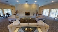 This single-floor space centers around a striking lounge area defined by a commanding floor-to-ceiling stone fireplace clad in large-format travertine or limestone tiles, serving as the room's focal point. The neutral, earthy tones of the stonework complement the warm beige and cream color palette found throughout the space. Recessed lighting in the ceiling provides ambient illumination, enhancing the room's welcoming atmosphere.

The seating arrangement is thoughtfully curated, featuring plush cream-toned sofas and armchairs arranged symmetrically around two glass-and-metal coffee tables, promoting both comfort and conversation. A richly patterned area rug in deep brown anchors the central seating area, adding texture and visual depth to the room.

Decorative accents, including tall floral arrangements in rich autumnal hues, vases, and sculptural objects, add personality and warmth to the space. Natural light filters in through a window or door visible to the right, contributing to the bright and airy feel of the interior. The overall ambiance is sophisticated yet comfortable, making it well-suited for relaxed gatherings or quiet retreats.