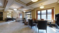 Located at 224 1st Ave in Columbus, Ohio's Short North area, this single-floor space presents a refined and welcoming lobby environment with notable architectural character. The ceiling is a standout feature, showcasing coffered detailing with gold-toned trim and recessed molding that adds a sense of grandeur and historical elegance. Pendant light fixtures hang at varying heights, complementing the natural light that pours in through large windows dressed with warm, rust-colored drapery, offering views of the surrounding urban landscape.

The flooring consists of large-format stone tiles with a subtle geometric border inlay, contributing to the space's polished, upscale aesthetic. A reception desk anchored near the center serves as a functional focal point, flanked by a seating area furnished with dark leather armchairs and studded upholstery details, alongside a built-in bench with decorative cushions. Decorative columns and paneled walls reinforce the classical architectural language throughout. The overall ambiance is professional yet inviting, blending historic design elements with contemporary comfort, making it well-suited as a hotel lobby or corporate reception area.
