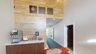 The lobby of Aspen Lofts makes an immediate impression through its thoughtfully designed interior. The focal point is a tall, light-toned wood-paneled accent wall that draws the eye upward, lending the space a sense of generous vertical height. Prominently displayed on this wall are the Aspen Lofts logo and branded signage, reinforcing the identity of the property with a clean, modern aesthetic.

To one side, a built-in coffee bar station features dark cabinetry topped with a light countertop, stocked with cups and small accessories, offering a welcoming amenity for residents and guests. The flooring at the entrance is a standout design element, composed of bold geometric shapes in red, orange, and neutral tones that create an energetic, contemporary feel.

A hallway extending from the lobby is finished with clean white walls and carpeted in a deep teal, guiding occupants further into the building. Recessed lighting throughout provides consistent, even illumination, while the overall color palette—warm wood tones, crisp whites, and vivid floor accents—creates an inviting and polished atmosphere. The single-story layout contributes to an accessible, cohesive flow throughout the space.