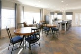 This single-floor gathering space is thoughtfully designed to offer both comfort and functionality in a welcoming, communal setting. The open layout seamlessly connects a dining area, lounge seating, and a kitchenette, creating a cohesive environment ideal for socializing and events.

The main gathering area is anchored by a striking fireplace with a dark stone surround and a wall-mounted flat-screen television above it, framed by crisp white wainscoting that lines the walls throughout. Recessed lighting and crown molding add a polished, refined touch to the neutral gray walls. The space features distinctive slate tile flooring with rich, earthy tones that lend character and warmth underfoot.

A round reclaimed wood dining table with a chevron-patterned top serves as a central focal point, surrounded by black metal chairs. A long bar-height counter with dark granite and seating for multiple guests provides an additional gathering spot. The lounge area includes plush brown leather armchairs accented with patterned throw pillows, positioned to take full advantage of the large windows that flood the room with natural light.

The kitchenette is well-appointed with white cabinetry, dark granite countertops, a double sink, a microwave, and patterned valances that add a decorative accent. Generous windows throughout the space offer views of the surrounding exterior, enhancing the bright and airy ambiance.