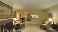 This single-floor residence features an open-concept layout that connects the living area, dining nook, and kitchen in one cohesive space. The living area is carpeted and generously proportioned, offering ample room for multiple seating arrangements. Warm beige walls and a textured ceiling contribute to the cozy, neutral ambiance throughout.

The kitchen is positioned at the far end of the main space and is accessible via a pass-through counter, which opens visibility between the cooking and living areas. White cabinetry and appliances, including a microwave, are visible from the main room, lending a clean and functional aesthetic to the kitchen zone.

Adjacent to the kitchen, a small dining nook accommodates a compact table and chairs, making it practical for everyday meals. A decorative metal shelving unit nearby adds a touch of character to the transitional space.

The living area is anchored by a cushioned sofa and accent chairs with patterned upholstery, complemented by a side table with a table lamp that adds warm ambient lighting. Framed artwork on the walls enhances the finished feel of the interior. A closeted entry area is also visible near the front of the space.