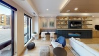 This thoughtfully designed lounge spans a single floor and is organized into several distinct zones that together create a versatile and inviting communal environment. The interior is anchored by light hardwood flooring throughout, which lends a warm, cohesive feel to the entire space.

A central social area features a comfortable arrangement of sofas, accent chairs, and ottomans positioned around a low coffee table, complemented by a striking built-in media wall. The media wall combines a mounted television with open wood shelving displaying books and décor, framed by deep navy tile detailing that adds a bold design contrast.

Adjacent to this area, a row of high-back upholstered booth seating lines one wall, each booth fitted with individual pendant lighting and a small table, offering semi-private nooks ideal for focused conversations or quiet work. A wood-paneled ceiling feature above adds architectural warmth to this section.

A dedicated workspace area includes a worktable with seating, office equipment, and wall-mounted magazine displays, providing practical functionality. Large windows along the perimeter allow natural light to filter in while offering views to the surrounding exterior. The overall ambiance is modern, welcoming, and well-suited for both collaboration and relaxation.