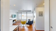 Located on Centre Street South in Calgary, this thoughtfully designed one-bedroom, one-bathroom unit offers a bright and contemporary living experience with sweeping city views. The open-plan layout connects the living area and kitchen seamlessly, making efficient use of the well-proportioned interior. Rich hardwood flooring runs throughout the main living spaces, adding warmth to the clean, white aesthetic.

The living room is a standout, featuring expansive floor-to-ceiling windows that frame panoramic views of the Calgary skyline and surrounding cityscape. A wall-mounted TV, a dedicated workspace with a built-in desk, and modern furnishings — including a vibrant yellow accent chair — give the space a stylish, functional character. The integrated kitchen features sleek white cabinetry, stainless steel appliances, and a cooktop, all finished to a high standard.

The bedroom is a serene retreat, with large windows showcasing striking views of the iconic Bow Tower's geometric façade. Sliding panel doors provide clean, minimalist access to the closet space. A wall-mounted TV adds convenience to the room.

The bathroom is finished in elegant marble-effect tiling from floor to ceiling, featuring a glass-enclosed walk-in shower, a soaking tub, a floating vanity, and recessed lighting — creating a polished, spa-like atmosphere throughout.