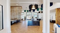 This thoughtfully designed single-floor office entrance spans a generous open layout that immediately impresses with its soaring coffered ceilings and walls of floor-to-ceiling windows that flood the interior with natural light. The entrance is anchored by a striking dark reception desk positioned beneath a dramatic arrangement of sculptural, layered pendant light fixtures in teal, navy, and black tones that serve as bold focal points throughout the space.

The flooring features a light-toned herringbone hardwood pattern that adds warmth and visual sophistication to the bright, white-walled interior. A tall natural wood slat partition wall adds organic texture and serves as an elegant divider between the open reception area and dedicated workstation zones, where multiple desks with leather seating accommodate staff and visitors alike.

A relaxed lounge area is furnished with white upholstered sofas accented with blue throw pillows, positioned near a sweeping watercolor-style ocean mural that brings an artistic, coastal element to the space. An octagonal mirror and console table provide additional decorative detail near a secondary entrance. Large glazed doors at the front entry offer clear views to an outdoor area with trees and covered outdoor seating, reinforcing a bright and welcoming ambiance throughout this well-appointed, professionally designed space.