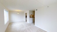 This single-floor space presents a clean, minimalist layout centered around a bright, open living area finished with light-toned tile or laminate flooring and crisp white walls that enhance the sense of openness. A simple pendant light fixture hangs from the textured white ceiling, providing a focal point and ambient illumination to the main living space. A centrally positioned partial wall divides the layout, creating a natural separation between zones while maintaining an open feel throughout.

To one side, a pass-through opening connects the living area to the kitchen, which features warm wood-toned cabinetry and stainless or light-colored appliances, including a refrigerator visible from the main space. This pass-through design promotes connectivity between the kitchen and living areas. A ceiling-mounted HVAC vent is also visible, indicating a ducted climate control system.

A secondary room, accessible through a doorway in the background, appears to serve as an additional living or utility space, also finished with white cabinetry and consistent neutral tones. The overall ambiance is light, airy, and neutral, offering a clean canvas adaptable to a variety of interior styles.