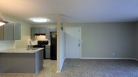 This single-floor unit offers an open-concept layout that connects the kitchen and living area in a practical, cohesive design. The kitchen is equipped with black appliances, including a refrigerator and electric range, complemented by grey cabinetry that provides ample storage. A light-toned countertop with a built-in sink extends along the kitchen perimeter, with a partial peninsula that subtly separates the cooking space from the rest of the unit. The kitchen features wood-style flooring, while the adjacent living area transitions to neutral carpet, adding a layer of comfort underfoot. A structural column near the entryway adds architectural definition to the open floor plan. The front door area is clean and unobstructed, offering a welcoming entry point into the space. Flush-mounted ceiling lighting provides consistent illumination throughout. The neutral wall tones in a soft sage-grey palette create a calm, cohesive atmosphere that ties the kitchen and living zones together. Overall, the space is compact yet well-organized, with a straightforward layout designed for efficient everyday living.