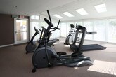 Located at 4133 Shoreline Cir in Virginia Beach, Virginia, this fitness center offers a bright and functional single-floor workout space designed to support a well-rounded cardio routine. The room is generously illuminated by a series of skylights set into the white paneled ceiling, complemented by large windows fitted with blinds along multiple walls, allowing natural light to fill the space throughout the day.

The fitness room is carpeted throughout and features a comprehensive selection of cardio equipment, including treadmills, elliptical trainers, upright and stationary cycling bikes, and a rowing machine. The variety of machines ensures users have multiple options for low-impact and high-intensity cardiovascular exercise. A wall-mounted television is positioned for convenient viewing during workouts.

The interior design blends two distinct wall treatments — warm taupe painted drywall on one side and white horizontal-paneled walls on the other — creating a clean, welcoming atmosphere. A wall-mounted mini-split HVAC unit ensures climate control year-round. An exterior-facing door provides direct access to the outdoors, adding convenience and ventilation options. The layout is open and spacious, providing ample room to move between equipment comfortably. Overall, this fitness center delivers a well-lit, fully equipped environment ideal for daily cardio exercise.