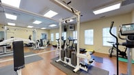 This dedicated fitness center offers a thoughtfully designed workout environment that combines functionality with a clean, open aesthetic. The space features warm hardwood flooring throughout, complemented by protective rubber fitness mats positioned beneath the equipment areas for safety and stability. Large windows along the walls invite generous natural light, creating a bright and energizing atmosphere that enhances the overall workout experience. Recessed and flush-mounted ceiling light panels provide additional illumination throughout the room. A prominent cable weight machine serves as a centerpiece of the strength training area, accompanied by additional resistance equipment and benches to support a variety of exercises. Cardio equipment, including a treadmill and stationary bike, is positioned along the perimeter of the room. Full-length mirrors line one wall, amplifying the sense of space and allowing users to monitor their form during workouts. The neutral, warm-toned walls contribute to the clean and welcoming ambiance of the space. The layout is well-organized, allowing for comfortable movement between equipment stations, making this fitness center a practical and inviting space for a comprehensive exercise routine.