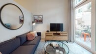 This single-floor residence offers a thoughtfully designed layout that integrates living, dining, sleeping, and bathroom spaces into a cohesive and functional environment. The kitchen is outfitted with stainless steel appliances, including a range, built-in microwave, and dishwasher, complemented by white cabinetry extending to the ceiling, a tile backsplash with under-cabinet lighting, and light hardwood-style flooring. A rustic wood dining table adjacent to the kitchen provides a practical and warm contrast to the modern finishes.

The living area features neutral walls and light flooring, with natural light filtering through floor-to-ceiling curtains. A charcoal sectional sofa, glass-top coffee table, and a wood media console create a relaxed yet contemporary atmosphere, enhanced by curated wall art and a round accent mirror.

The bedroom maintains a calm, neutral palette with carpeted flooring, a upholstered queen bed flanked by walnut-toned nightstands with sleek task lamps, and a vibrant abstract painting as a focal point above the headboard.

The bathroom is finished with a white quartz countertop, undermount sink, brushed gold fixtures, a large frameless mirror, and open shelving for organized storage, contributing to an overall clean and modern aesthetic.