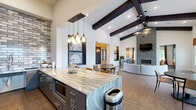 Located at 13872 Del Corso Way in Broomfield, Colorado, this thoughtfully designed single-floor community space blends modern sophistication with inviting warmth. The open-concept layout is anchored by dramatic soaring ceilings adorned with bold dark wood exposed beams, creating a striking architectural statement that carries throughout the entire space. Wide-plank wood-look tile flooring unifies each area seamlessly.

The living area centers around a floor-to-ceiling stacked stone fireplace, topped with a wall-mounted television, and is furnished with a curved sectional sofa and stylish accent chairs, offering a comfortable gathering space. Abundant natural light pours in through large windows dressed with warm-toned roman shades.

The kitchen is outfitted with sleek slate-grey cabinetry, sweeping granite countertops, stainless steel appliances, and a full brick-tile accent wall that adds industrial charm. Pendant lighting above the expansive island enhances both function and style.

The dining area features multiple round tables with modern seating beneath recessed lighting and curated wall art set within illuminated niches. A dedicated workspace nook includes built-in desks set against a bold geometric accent wall, ideal for productivity. Together, these spaces form a cohesive, upscale environment designed for both socializing and focused work.