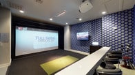 The Golf Room is a purpose-built indoor golf simulation space designed to deliver an immersive and entertaining experience for players of all skill levels. At the heart of the room is a Full Swing Simulator, complete with a large high-definition projection screen and overhead projector, offering a variety of play, practice, and event modes. A green artificial turf hitting mat is integrated into the carpeted floor, creating a realistic feel for each swing. The room's interior is defined by striking blue and silver hexagonal patterned wallcovering that lines multiple walls, lending a modern, dynamic aesthetic to the space. A wall-mounted flat-screen television and a dark wood media cabinet provide additional viewing and control options. Along one side of the room, a sleek white high-top bar counter is lined with adjustable chrome and black bar stools, offering comfortable seating for spectators and guests. Recessed ceiling lighting illuminates the space evenly, complementing the dark tones of the simulator bay. The room is accessed through a glass-paneled door and features clean transitions between the tiled entryway and the carpeted simulator area, creating a polished and well-appointed environment ideal for golf enthusiasts.