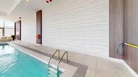 Located at 180 Metcalfe Street in Ottawa, this sophisticated indoor aquatic amenity space offers a refined retreat designed with modern elegance. The centerpiece is a generously sized lap pool featuring crystal-clear turquoise water, framed by smooth light-toned porcelain tile surrounds and stainless steel entry rails for safe and easy access. Adjacent to the pool sits a dedicated hot tub, providing a relaxing complement to the main swimming area.

The space is architecturally striking, with floor-to-ceiling windows supported by substantial concrete columns along one full side of the room, flooding the interior with abundant natural light while offering expansive views of the Ottawa skyline. A comfortable lounge area with plush, curved neutral-toned seating and lush potted greenery is positioned poolside, creating an inviting space to relax and take in the panoramic views.

The interior finishes are polished and contemporary, featuring textured white stone-effect wall cladding on one end and rich warm wood paneling on the other, adding depth and contrast to the bright, airy atmosphere. Recessed ceiling lighting complements the natural light throughout. Safety equipment is readily accessible, and a glass-paneled entry door provides a seamless transition into the facility. The overall ambiance is calm, upscale, and wellness-focused.
