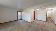 This single-floor space offers a straightforward, functional layout designed for comfortable everyday living. The main living area is generously proportioned, featuring neutral wall-to-wall carpeting, crisp white walls, and warm wood-toned baseboards and door trim throughout. A door with a windowed panel allows natural light to filter into the space, while multiple electrical outlets are conveniently positioned along the walls.

Adjacent to the living area, a distinct dining space is defined by tile flooring and illuminated by a globe pendant light, creating a subtle separation between the two zones. A door entry nearby provides additional access to the unit.

The bedroom is a comfortable, well-sized room with plush carpeting, a large window fitted with vertical blinds that invite natural light, and a textured ceiling. The room offers ample wall space suitable for a variety of furniture arrangements.

The full bathroom features classic small-format white tile flooring, a wood-cabinet vanity with a white countertop and integrated sink, a toilet, and a shower-tub combination enclosed by a decorative curtain. A textured ceiling and built-in ventilation complete the space.

Overall, the unit presents a clean, neutral palette with functional finishes throughout.