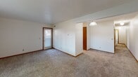 This single-floor space offers a straightforward, functional layout designed for comfortable everyday living. The main living area is generously proportioned, featuring neutral wall-to-wall carpeting, crisp white walls, and warm wood-toned baseboards and door trim throughout. A door with a windowed panel allows natural light to filter into the space, while multiple electrical outlets are conveniently positioned along the walls.

Adjacent to the living area, a distinct dining space is defined by tile flooring and illuminated by a globe pendant light, creating a subtle separation between the two zones. A door entry nearby provides additional access to the unit.

The bedroom is a comfortable, well-sized room with plush carpeting, a large window fitted with vertical blinds that invite natural light, and a textured ceiling. The room offers ample wall space suitable for a variety of furniture arrangements.

The full bathroom features classic small-format white tile flooring, a wood-cabinet vanity with a white countertop and integrated sink, a toilet, and a shower-tub combination enclosed by a decorative curtain. A textured ceiling and built-in ventilation complete the space.

Overall, the unit presents a clean, neutral palette with functional finishes throughout.