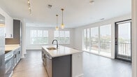 This well-designed two-bedroom, two-bathroom residence offers a thoughtfully arranged single-floor layout with a bright, open-concept living space at its core. The kitchen serves as a central focal point, anchored by a large island with a white quartz countertop, a matte black faucet, and an undermount sink. Two-toned cabinetry — combining white upper cabinets with slate gray lower units — pairs with stainless steel appliances, subway tile backsplash, and dual brass pendant lights to create a polished, modern aesthetic. Wood-style plank flooring flows seamlessly through the main living area, which is lined with expansive windows and a glass sliding door, flooding the space with natural light and offering views of the surrounding cityscape.

A hallway connects the living area to both bedrooms, each finished with neutral carpet, ceiling fans, and natural light from windows. The primary bedroom features floor-to-ceiling windows and direct access to a well-appointed en-suite bathroom, which includes gray cabinetry, a white countertop, dual sinks with matte black hardware, and penny-tile flooring. Two closets — including a spacious walk-in — provide ample storage throughout the residence.