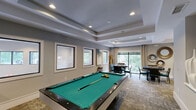 Located at 131 Poindexter Dr in Charlotte, NC, this thoughtfully designed single-floor amenity space offers a seamless blend of comfort and entertainment. The layout is divided into two well-appointed areas: a living room and a game room, together forming a cohesive and inviting environment.

The living room is anchored by a large L-shaped sectional sofa in neutral cream tones, accented with richly colored throw pillows in burnt orange and teal. A glass-top coffee table and a sleek media console with sculptural decorative pieces add refined touches. A wall-mounted flat-screen TV is positioned for optimal viewing, while a bold abstract artwork provides a vibrant focal point. Tray ceilings with recessed lighting lend architectural depth, and expansive windows dressed with floor-length drapes invite generous natural light and serene greenery views.

The game room is centered around a striking billiard table with a green felt surface, set against a backdrop of a moss-framed wall-mounted TV flanked by tall white vases. Multiple windows and glass-panel doors fill the space with daylight, while tray ceilings echo the refined architectural language found throughout. A casual seating area with round tables and upholstered chairs offers a comfortable space for card games or socializing, complemented by decorative wall mirrors and curated artwork.