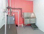 This single-floor space is purpose-built for professional pet grooming, offering a functional and well-equipped environment. The room features concrete block walls painted in a light mint green on two sides and a bold coral-orange accent wall, lending a clean, utilitarian aesthetic with a touch of color. A drop ceiling with recessed lighting provides consistent overhead illumination throughout the space.

The grooming area is outfitted with a height-adjustable grooming table fitted with an overhead arm and hook for securing animals during grooming sessions. Along the far wall, a stainless steel bathing station with a step-up ramp provides a hygienic and accessible wash area, ideal for animals of varying sizes. A large water heater is centrally positioned and connected to visible plumbing lines routed along the walls, ensuring a reliable hot water supply for bathing needs.

Exposed piping in black and red runs along the coral accent wall, reflecting the room's practical, no-frills design. The concrete floor is easy to clean and maintain, making it well-suited for the demands of a professional grooming environment. Overall, the space is efficiently organized to support a streamlined grooming workflow.