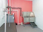 This single-floor space is purpose-built for professional pet grooming, offering a functional and well-equipped environment. The room features concrete block walls painted in a light mint green on two sides and a bold coral-orange accent wall, lending a clean, utilitarian aesthetic with a touch of color. A drop ceiling with recessed lighting provides consistent overhead illumination throughout the space.

The grooming area is outfitted with a height-adjustable grooming table fitted with an overhead arm and hook for securing animals during grooming sessions. Along the far wall, a stainless steel bathing station with a step-up ramp provides a hygienic and accessible wash area, ideal for animals of varying sizes. A large water heater is centrally positioned and connected to visible plumbing lines routed along the walls, ensuring a reliable hot water supply for bathing needs.

Exposed piping in black and red runs along the coral accent wall, reflecting the room's practical, no-frills design. The concrete floor is easy to clean and maintain, making it well-suited for the demands of a professional grooming environment. Overall, the space is efficiently organized to support a streamlined grooming workflow.