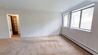 This single-floor, 2-bedroom, 1-bathroom unit offers a straightforward and functional layout designed for comfortable everyday living. The main living area features warm, neutral-toned carpet underfoot, crisp white walls, and a half-wall partition that separates the space while maintaining an open feel. A closet near the entryway provides convenient storage, and natural light filters in through windows that overlook the surrounding exterior. The kitchen is efficiently arranged in a galley style, lined with honey-toned oak cabinetry on both sides offering ample storage. Light countertops, a stainless steel double sink, and white appliances including a refrigerator and electric range complete the space, all set on easy-to-clean tile flooring. Both bedrooms are carpeted and feature white walls with baseboard heating units running along the perimeter, ensuring warmth throughout the colder months. One bedroom includes a window that brings in natural light. The full bathroom is clean and simply appointed, featuring a tiled tub-shower combination, a toilet, and tile flooring. Grab bars are installed for added accessibility. The unit's consistent neutral palette and practical design make it a versatile and move-in ready home.