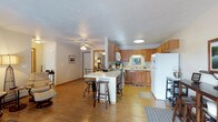 This single-floor residence offers a practical and welcoming layout centered around an open-concept living and kitchen area. The space is unified by warm hardwood flooring that extends throughout, creating a cohesive and inviting atmosphere.

The kitchen is well-appointed with classic honey-toned wood cabinetry offering generous storage, a white refrigerator, and a range with overhead microwave. A white breakfast bar serves as the centerpiece of the space, providing casual seating with bar-height stools — perfect for dining or entertaining. The kitchen also features a sink positioned beneath a window, allowing for natural light to brighten the workspace.

A ceiling fan with integrated lighting is installed in the adjoining living area, providing both comfort and illumination. The open sightlines between the kitchen and living area create a spacious, airy feel, making the floor plan well-suited for everyday living and hosting guests.

A hallway visible from the main living area leads to additional rooms, suggesting a thoughtfully organized layout that balances shared and private spaces. The overall design is functional and homey, with neutral tones, warm wood finishes, and practical fixtures that contribute to a comfortable, lived-in ambiance throughout the home.