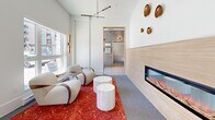Located at 1110 McCallum Rd in Langford, British Columbia, this thoughtfully designed building lobby offers a polished and welcoming first impression. The space is organized across a single level and comprises a central living area complemented by three connecting hallways, creating a logical and intuitive flow throughout.

The entry foyer is finished with large-format light grey tiles and opens through floor-to-ceiling glass double doors, allowing natural light to pour in and providing clear sightlines to the street. Stainless steel elevator doors are conveniently positioned just off the entrance, framed by clean white walls and warm wood-toned accents.

The living area is a standout feature, boasting soaring vaulted ceilings that lend a sense of spaciousness. A sleek, linear fireplace set within a textured wood-panel surround serves as the room's focal point, while sculptural bronze wall decorations and a modern multi-arm chandelier add artistic character. Curved, upholstered lounge chairs arranged around cylindrical stone side tables invite residents and guests to relax, anchored by a rich, patterned red area rug over grey carpeting.

Large windows frame views of the surrounding neighbourhood, filling the space with abundant natural light. The hallways feature large-format grey tile walls and flooring, maintaining a cohesive and contemporary aesthetic throughout this refined communal space.